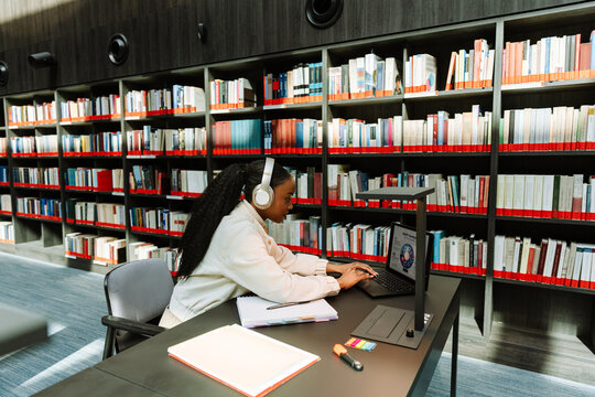 Female student typing on laptop and sitting at desk while listening to headphones
