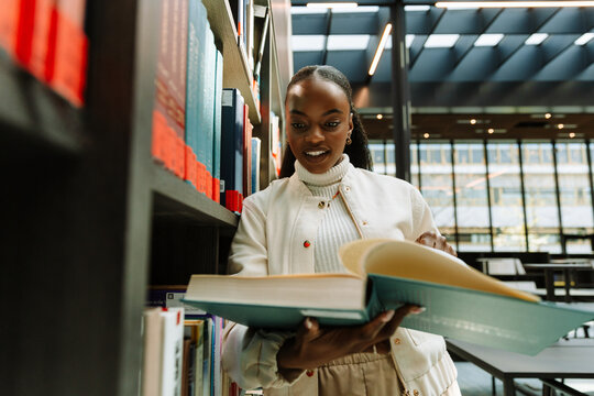 Female student holding a book she is reading and leaning on bookshelves - Powered by Adobe