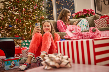 Two girls in matching pajamas relax on couch surrounded by Christmas tree and wrapped presents
