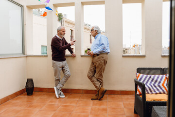 Fototapeta premium A man talks to a male friend who listens as they stand and lean on a windowsill while holding glasses