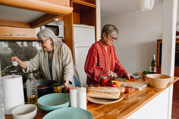 A woman is salting and talking while a female friend is standing next to her at the table and cutting carrots