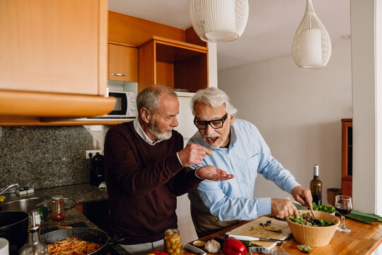 A man gives a taste of an olive to his male friend who stirs it in a bowl while they stand at the table