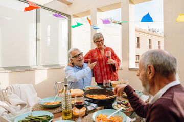 A woman stands and holds a glass while hugging a man who is talking and pointing at her while sitting at a table and they are smiling