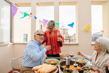 A woman and a man are talking and sitting at a table while he is hugged by a woman who is standing and holding a glass