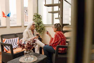 Two female friends holding glasses and sitting on chairs facing each other while one of them listens and laughs