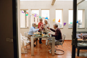 Two female friends are standing and one of them is talking while two male friends are sitting next to them at a table and they are holding glasses
