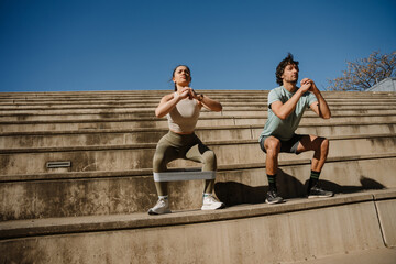 A woman and a man hold their hands together in front of each other and squat on the bleachers while she has elastic bands on her legs