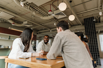 A male worker is talking and sitting at a table with a group of three workers while one of them is...
