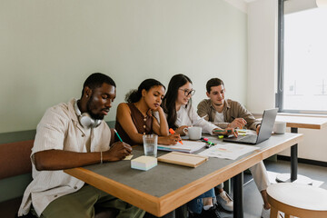 A group of four workers sit at a table while two of them write on sticky notes and a notebook and two of them smile and look at a laptop