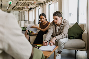 A female employee is holding documents and talking to a male employee who is sitting next to her on the couch and writing in a notebook