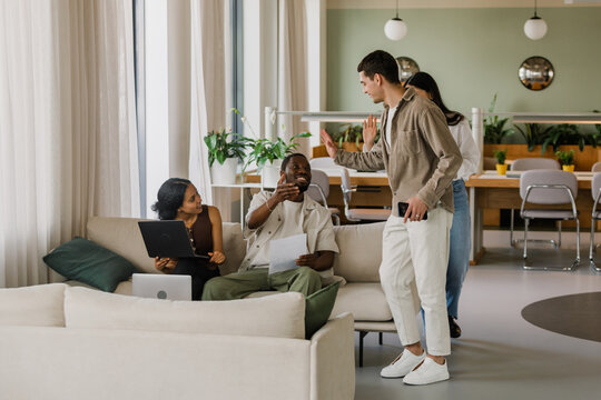 Male and female employees walk and hold hands to give high five to male and female employees sitting on the couch while they smile
