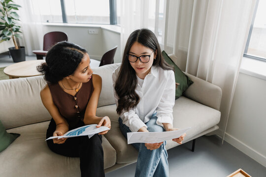Two female office workers talking and holding documents while sitting on a couch - Powered by Adobe