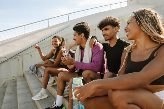 A group of five athletes are laughing and sitting on the stairs while two of them are holding bottles - Powered by Adobe
