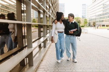 Male student laughing and holding notebooks while listening and hugging female student walking next to him and talking