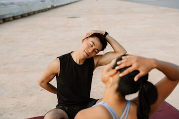 A man with his eyes closed sits on a mat opposite a woman while they stretch while placing their hands on their heads, which are tilted to the side
