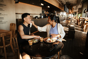 Female athlete wipes male athlete's face with a napkin while they laugh and sit at a table