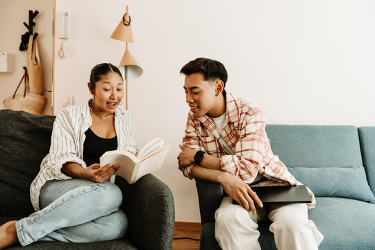 A man holds a laptop and sits on a couch while listening to a woman who is reading a book in surprise and sitting on a chair while they smile