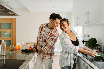 A man and a woman laugh and talk while she washes greens in the sink and he cuts tomatoes