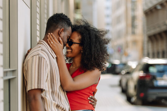 A woman laughs and puts her hand on the neck of a man who is leaning on a wall while they stand and hug - Powered by Adobe