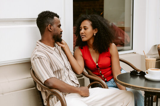 A woman touches a man's beard while they sit on chairs at a table and look at each other