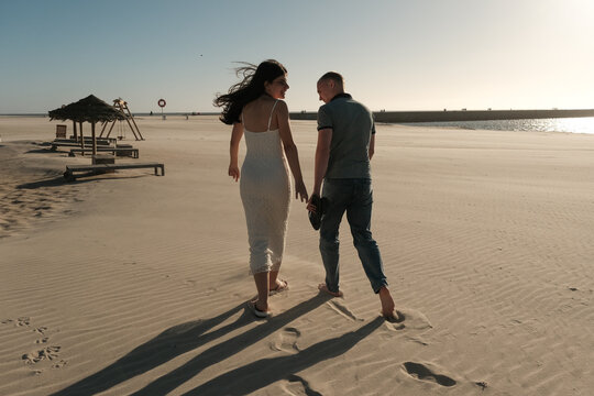Couple walking barefoot on sandy beach near ocean at sunset, wind blowing her hair in a peaceful romantic scene. - Powered by Adobe