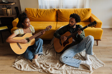 A male musician listens to a female musician singing while they play guitars and sit on a blanket while leaning on a sofa
