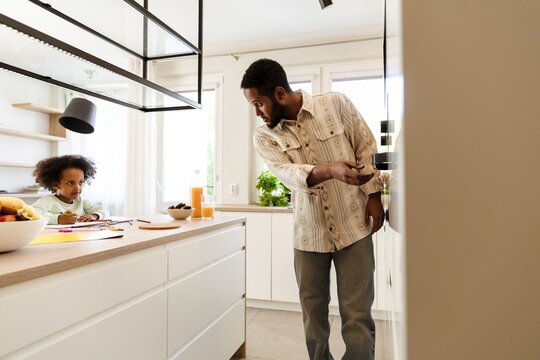 A father stands and holds his hand near the oven while talking to his daughter who sits at the table and listens while holding a pencil - Powered by Adobe