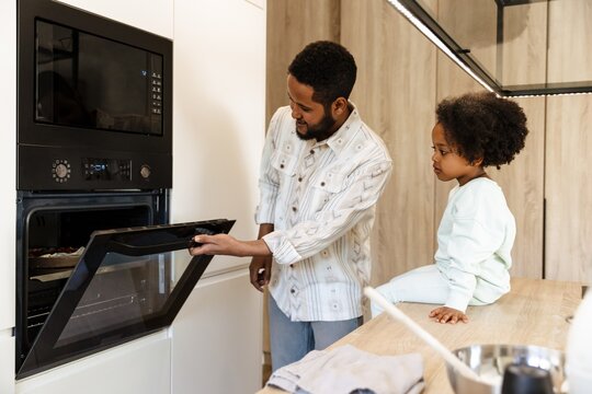 Daughter sits on the table and looks into the oven which her father opens while standing