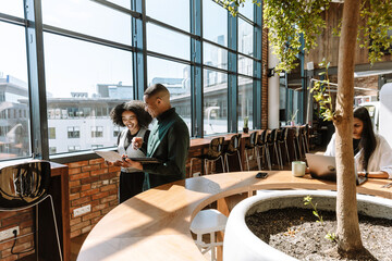 A male worker holds a computer tablet and laughs with a female worker walking next to him as they look at the documents she is showing