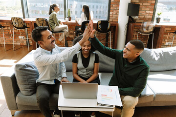Two male workers high-five each other while sitting on a couch with a female worker looking at a laptop and laughing