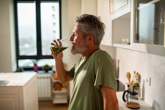 Mature man sipping green smoothie in home kitchen, content