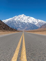 Naklejka premium A straight asphalt road with double yellow lines stretches towards a majestic snow-covered mountain range under a bright, clear blue sky. The landscape is arid