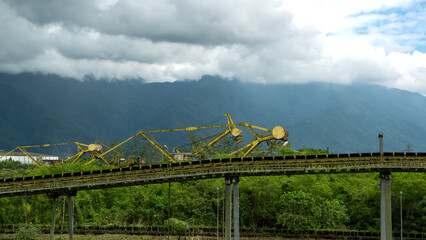 Fototapeta premium Old yellow industrial machinery and elevated conveyor system surrounded by dense greenery, set against misty mountains and heavy clouds