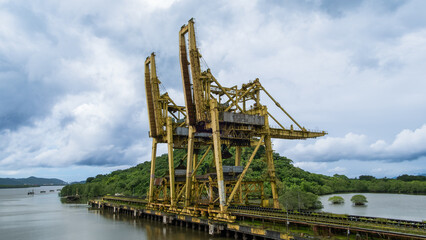 Two colossal abandoned cargo loading cranes on an industrial pier, surrounded by lush tropical mangroves under a dramatic cloudy sky