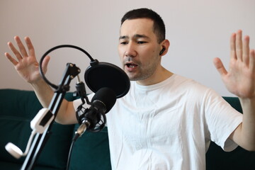 Asian man recording audio at home, speaking passionately into a microphone with raised hands. Podcasting, streaming, or voice-over session in a casual home studio