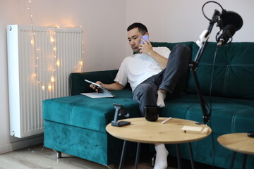 Asian man working from home on the sofa, multitasking with a phone call, tablet, and laptop. Casual remote-work setup with microphone, gadgets, and relaxed living-room environment