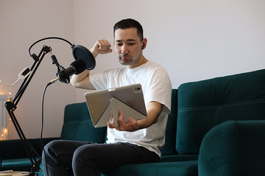 Asian man (Kazakh) recording a video call or online session at home, raising his hand in expression while speaking into a microphone and holding a tablet. Casual digital-creator setup