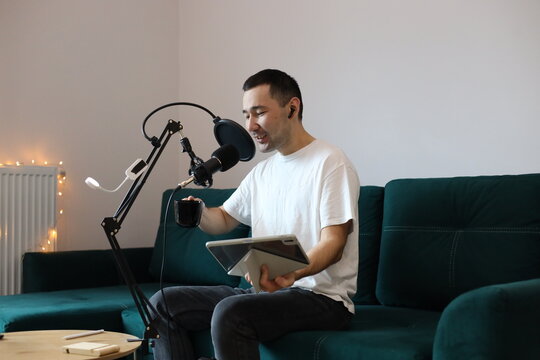 Asian man (Kazakh) recording audio or video at home, speaking into a microphone while holding a tablet and a coffee mug. Casual digital-creator workflow in a modern living space