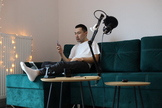 Asian man working from home on the sofa, multitasking with a phone call, tablet, and laptop. Casual remote-work setup with microphone, gadgets, and relaxed living-room environment