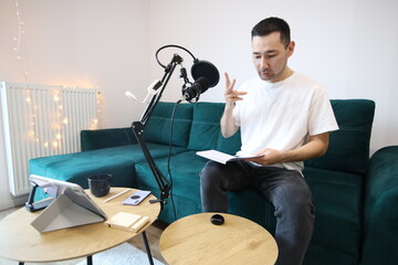 Asian man reviewing notes while recording audio at home, sitting by a microphone in a casual home studio. Focused remote-work or podcast preparation moment