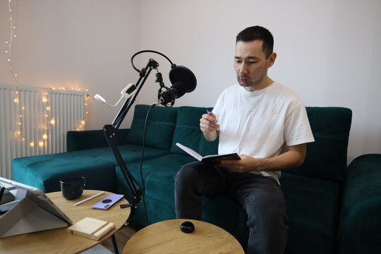 Asian man reviewing notes while recording audio at home, sitting by a microphone in a casual home studio. Focused remote-work or podcast preparation moment
