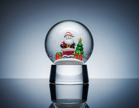 Santa claus figurine sitting in snow globe with christmas tree and gifts on reflective surface creating festive holiday spirit.