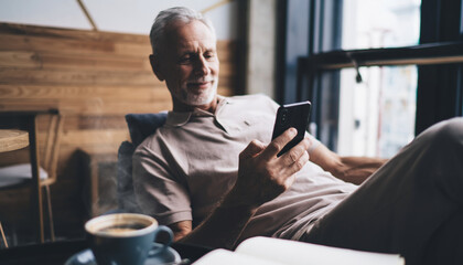 Senior man relaxing in cozy cafe, enjoying morning coffee while scrolling on smartphone, natural light, casual atmosphere, peaceful and content expression