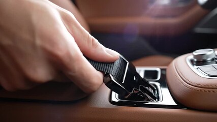 A hand holding a seatbelt buckle in a car with brown leather interior, preparing to fasten, showing upscale safety routine.
