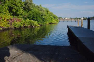 Tranquil Waterfront Dock Over Green bay With Reflective Water And Bright Sky Coffee Pot Park in St. Petersburg, FL. Wooden dock extending into clear water, lush green bushes along the bank, and a brig