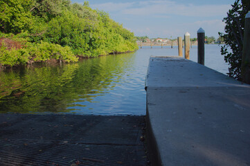 Tranquil Waterfront Dock Over Green bay With Reflective Water And Bright Sky Coffee Pot Park in St. Petersburg, FL. Wooden dock extending into clear water, lush green bushes along the bank, and a brig
