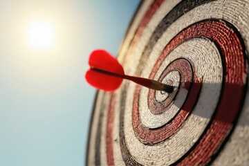 Red dart hitting bullseye on circular board against blue sky with bright sun, conveying determination and focus.