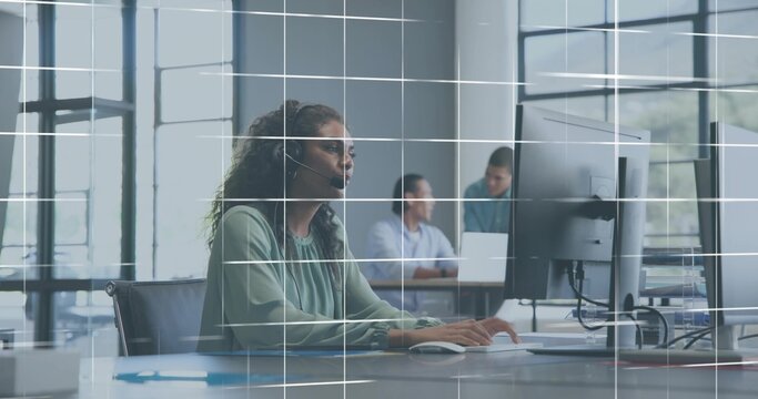 Typing woman in green blouse and headset using monitor at open-plan desk with laptop, glass grid - Powered by Adobe