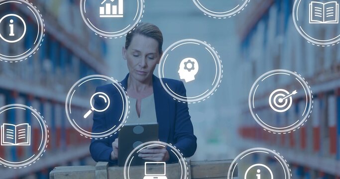 Standing mature woman holding tablet in warehouse aisle with pallet and icons, wearing navy blazer - Powered by Adobe
