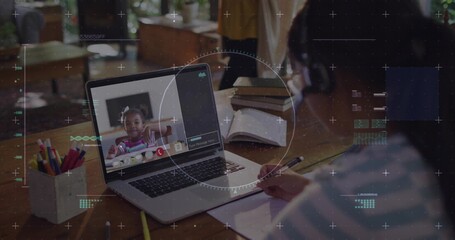 Writing Asian child in striped shirt looking at laptop on wooden table, notebook and pencil cup